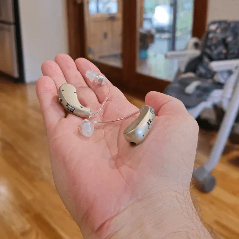 Two hearing aids held in a hand with a home interior background